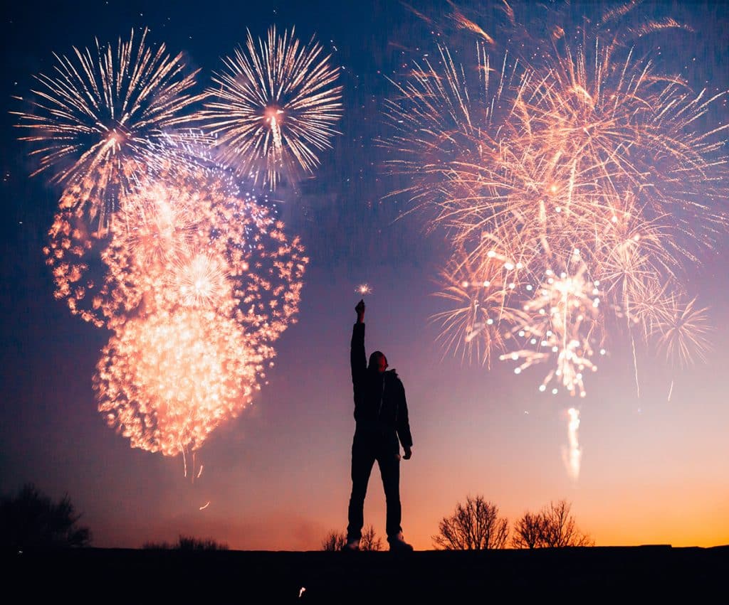 Eine Person, die auf erhöhtem Grund steht und eine Wunderkerze zum Himmel hält, während im Hintergrund Feuerwerk losgeht - Symbolbild für Silvester-Locations.