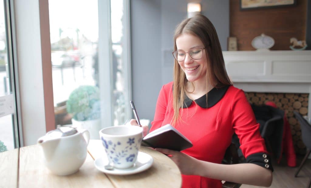 Eine junge Frau an einem Cafétisch lächelt und macht Notizen in einem kleinen Buch - Symbolbild für einen guten Wochenstart.
