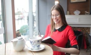 Eine junge Frau an einem Cafétisch lächelt und macht Notizen in einem kleinen Buch - Symbolbild für einen guten Wochenstart.