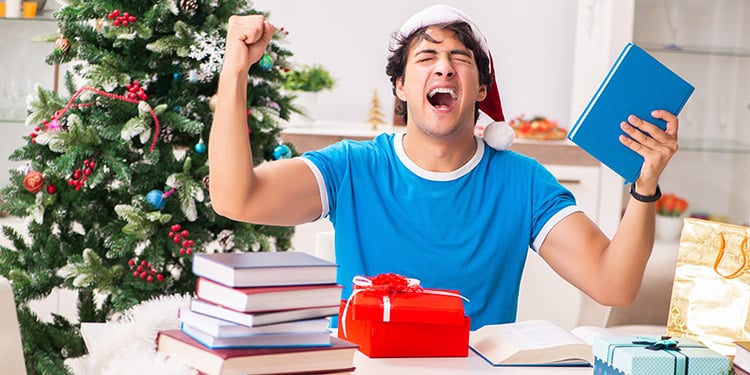 Ein junger Mann mit Buch in der Hand und Weihnachtsmütze auf dem Kopf sitzt an einem Tisch mit Büchern und Geschenken und jubelt mit erhobenen Armen. Im Hintergrund ein geschmückter Weihnachtsbaum - Symbolbild für DIY-Weihnachtsgeschenke.