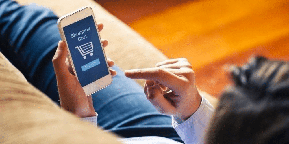 Eine Frau sitzt auf einem Sofa und legt auf ihrem Handy etwas in den Warenkorb - Symbolbild für Onlinebestellung