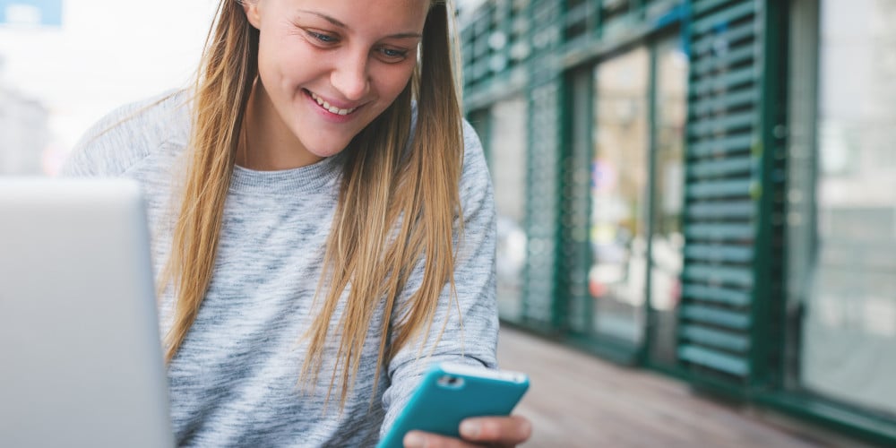 Blonde junge Frau schaut sitzt vor einem Laptop und schaut lächelnd auf ihr Smartphone.
